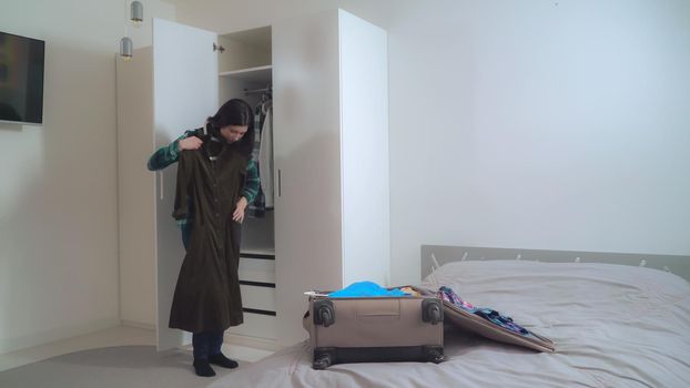 Lady try on dress and put it in suitcase. Young woman wearing in casual clothes packing bag planning vocation. Caucasian girl with black hair in apartment.