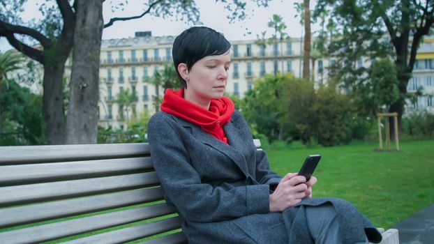 Female texting sms on the mobile phone. Caucasian model sits on the bench in the park. Wearing casual coat and red sweater. Outdoor lady portrait.