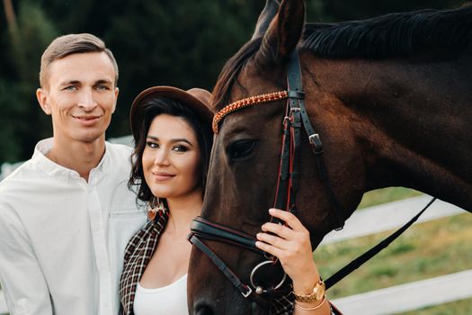 a pregnant girl in a hat and a man in white clothes stand next to horses near a white fence.Stylish pregnant woman with a man with horses.Married couple