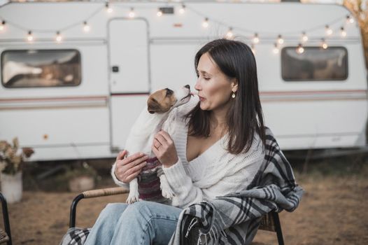 Caucasian woman sitting by a wagon in a wicker chair and hugging with Jack Russell Terrier dog outdoors. Travel in a camper in autumn