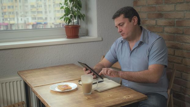 Adult male in a blue shirt in the morning sitting in the hotel drinking coffee and eating toast, is on the table and tablet daily, Soft panorama