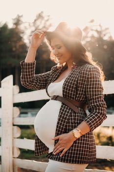 a pregnant girl with a big belly in a hat near a horse corral in nature at sunset.Stylish pregnant woman in a brown dress with horses