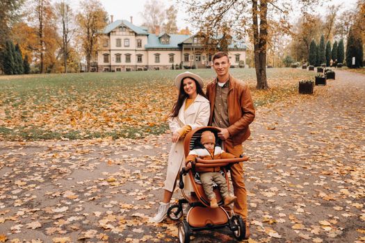 Father and mother on a walk with a child in a stroller in the autumn Park on the background of the estate. A family walks through the Golden autumn nature Park.