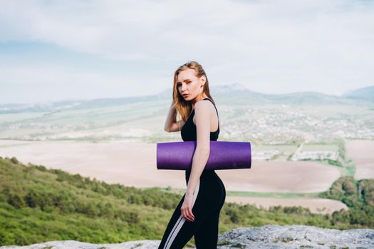 A girl athlete with a purple yoga mat under her armpit. A beautiful athlete in the mountains.