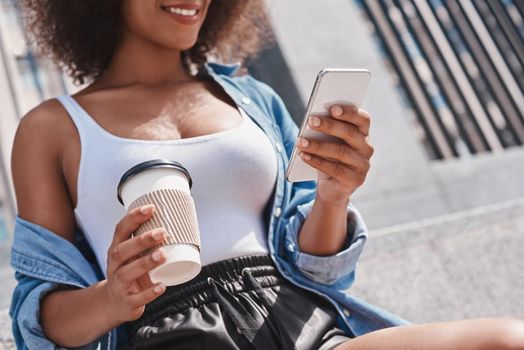 Young woman free style on the street sitting on concrete stairs holding cup of hot coffee browsing internet on smartphone smiling joyful close-up