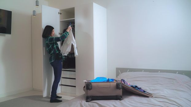 Young happy woman packing male and female clothes in suitcase. Smiling brunette planning traveling in apartment with casual interior.
