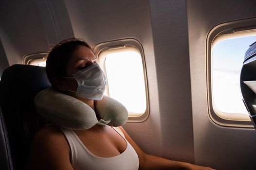 Caucasian woman on board aircraft in medical mask sleeping with neck pillow