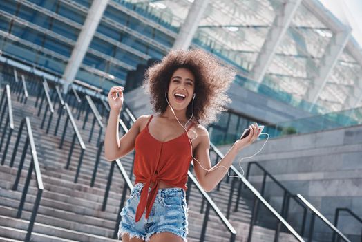 Young woman wearing earphones free style on the street jumping on concrete stairs holding smartphone listening to favorite song singing laughing cheerful