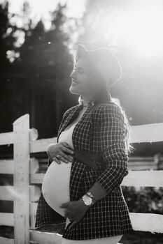 a pregnant girl with a big belly in a hat near a horse corral in nature at sunset.Stylish pregnant woman in a brown dress with horses.black and white photo.