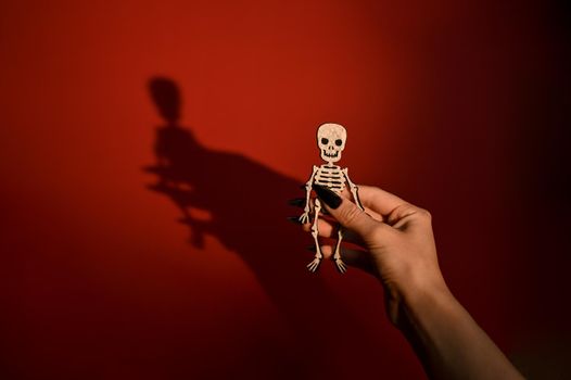 Postcard for Halloween. a human skeleton toy in hand with a shadow on a red background. Halloween symbol. copy space.