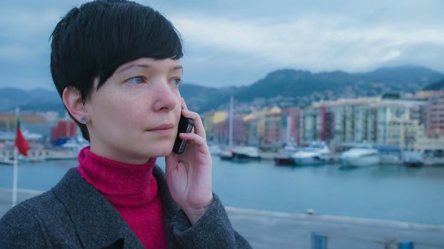 Elegant young female professional standing seafront has conversation on the mobile phone. On the background harbour with ships and yachts. Girl in winter season dressed casual grey coat.