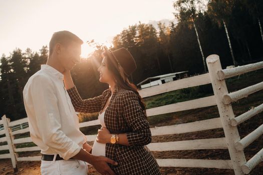 a pregnant girl in a hat and her husband in white clothes stand next to a horse corral at sunset.a stylish couple is waiting for a child in nature.