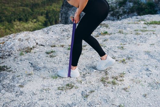 The girl does an exercise for the legs with the help of a rubber band. A beautiful athlete in the mountains.