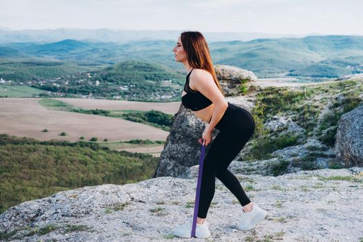 The girl does an exercise for the legs with the help of a rubber band. A beautiful athlete in the mountains.