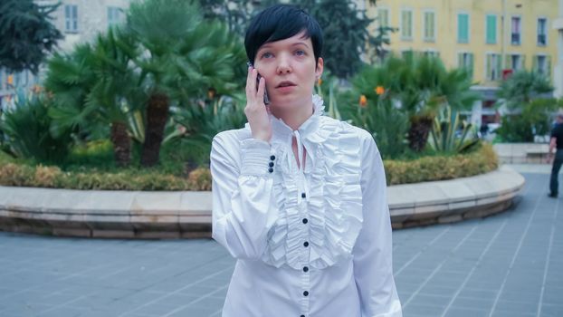 Attractive female in the square talking on the mobile phone. On The background road with cars, trees, shops and walking people. Caucasian model with short black hair dressed formal. Day time.