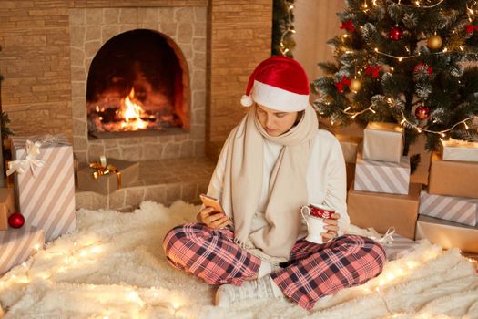 Photo of sick lady sit in festive living room using cellphone and hold beverage mug with christmas x-mas ornament, texting to somebody and congratulates with holidays.