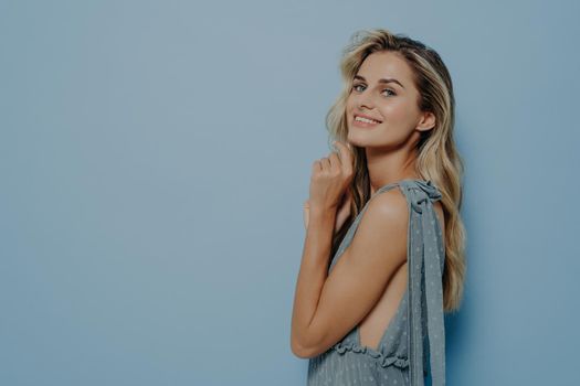 Curious sweet blonde woman in romantic blue dress with finger next to chin, looking at camera smiling with head tilted upwards, thinking about something good, standing isolated over blue wall