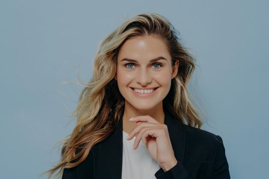 Studio shot of happy blonde girl with hand next to her chin, looking forward with bright smile, feeling calm relaxed while standing against blue background, dressed in black coat over white tshirt