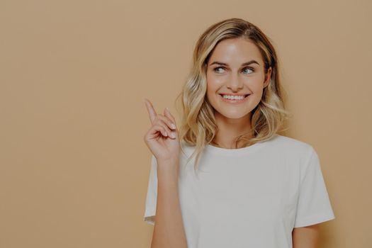 Look at that. Positive young woman customer with blonde hair wearing white tshirt with pleasant smile pointing at copy space on orange background. Advertising and promotion concept