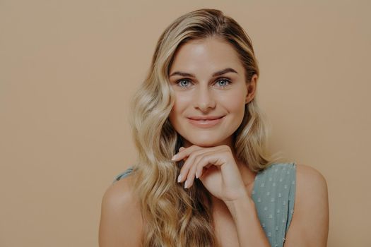 Beautiful relaxed young blonde woman in blue dress with hand under chin looking at camera with optimism, satisfaction and slight smile, standing isolated next to orange wall