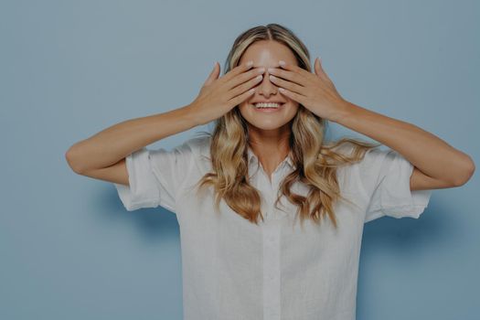Joyful blonde girl wearing white shirt covering her eyes with hands while smiling with toothy smile, does not want to see whats in front of her while standing against blue studio background