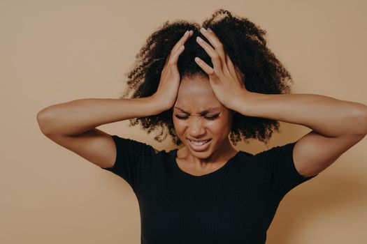 Studio shot of stressful and frustrated young african woman keeping head with both hands with closed eyes, displeased female suffering from headache or having problems at work. Stress and depression