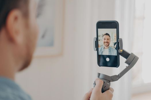 Handsome young man vlogging or recording video on smartphone with great handheld gimbal stabilizer, standing in living room at home background. Vlog and video blogging concept