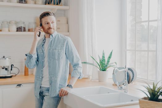 Young male in casual jeans shirt speaking on cellphone with work partner and looking away while standing by wooden table top in modern kitchen interior. Freelance and distance work concept