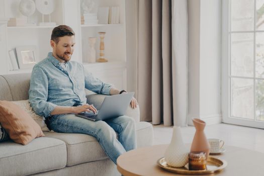 Young attractive smiling male freelancer sitting on comfortable beige sofa at home, using laptop and checking his emals while working remotely during weekend, wearing casual clothes