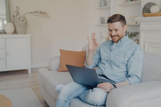 Young office male worker working remotely from home, showing ok gesture to his boss in online meeting while sitting on comfortable couch in modern living room with crossed leg