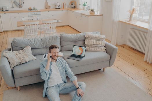 Young male bussiness owner receiving call from his worker, happy with his company profits, smiling after checking report on laptop, sitting on carpet while resting against sofa. Freelance concept