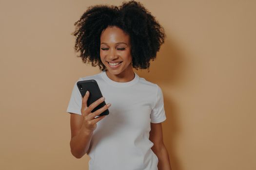 Happy smiling african female student using mobile phone and smiling, reading positive news in internet or chatting with boyfriend online, holding modern smartphone while posing against sand color wall
