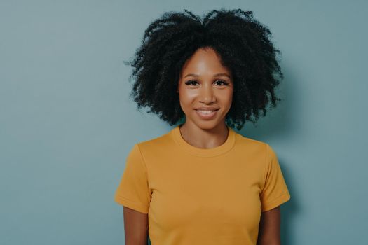 Waist up portrait of cheerful young mixed race woman with curly hair posing in studio with happy smile. Dark-skinned woman wears yellow tshirt, smiling cheerfully, showing her white straight teeth