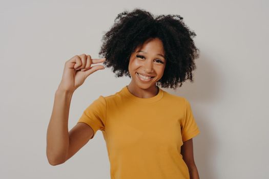Cheerful smiling African American woman gestures small size with fingers, asks for little bit time, or measures too small object, shows something minimal, dressed casually stands in studio background