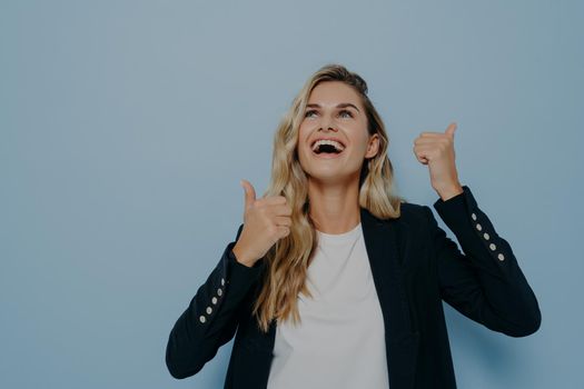 Bright laughing young girl slightly tilted her head back showing thumbs up ok gesture with both hands, happy to approve or to recommend something very good, posing isolated over blue studio wall