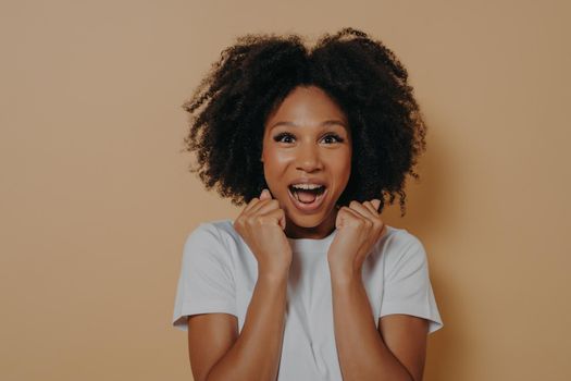 Portrait of excited amazed young woman keeping mouth opened and screaming with enjoyement, clenching fists, celebrating victory and success while posing against beige wall in studio. Triumph concept