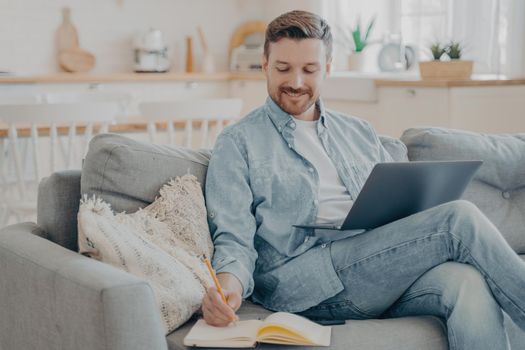 Young office worker working remotely from home, researching in internet using notebook, writing information down into red note book with pencil while sitting on cozy couch