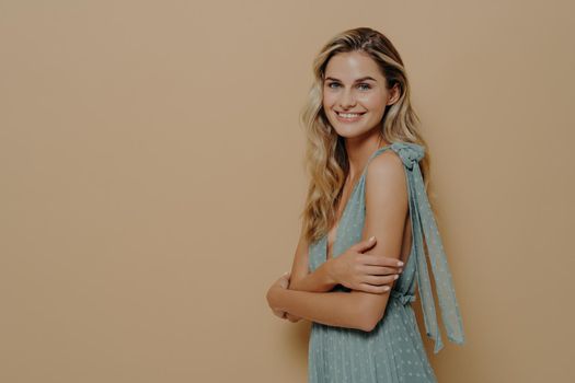 Romantic young and attractive female model in beauriful evening dress getting ready for perfomance standing sideways delighted and happy with crossed hands, smiling joyfully at camera