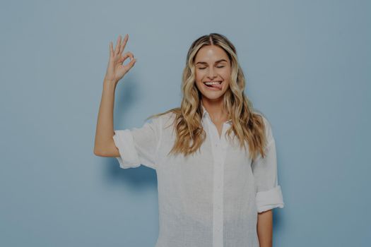Young joyful blonde caucasian woman demonstrating agreement and showing okay gesture with closed eyes, sticking out her tongue and making funny face expression. Body language concept