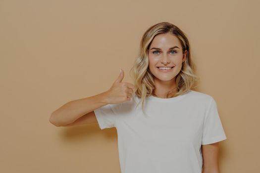 I like that. Happy young blonde female wearing white casual tshirt making thumbs up gesture and smiling cheerfully at camera, posing against yellow background. Body language concept