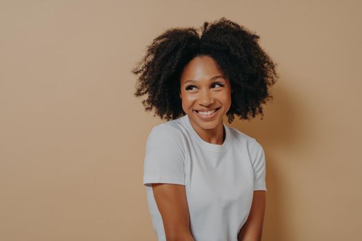 Waist up of cute curly african woman with toothy smile dressed in white t shirt looking aside with shyness and expressing positive emotions, isolated over beige background. Face expressions concept