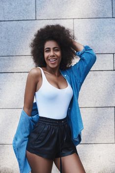 Young woman wearing jeans jacket free style on the street standing isolated on concrete wall touching hair looking camera laughing playful