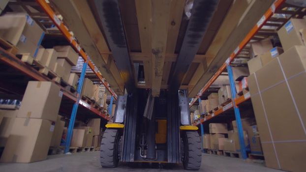 Worker drives forklift trucks with cardboard boxes in warehouse. Man rides along metal racks in the storehouse