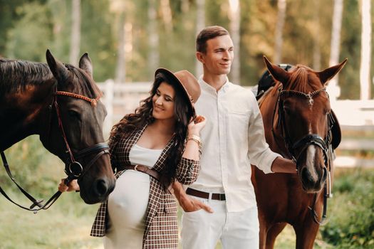 a pregnant girl in a hat and her husband in white clothes stand next to horses in the forest in nature.Stylish pregnant woman with a man with horses.Family