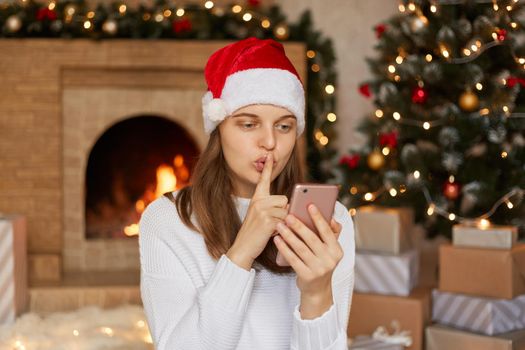 Woman in santa claus hat holding mobile phone in hands, looks at its screen, having video call, asks somebody too keep her secret, keeping finger near lips, lady wearing white sweater, posing indoor.