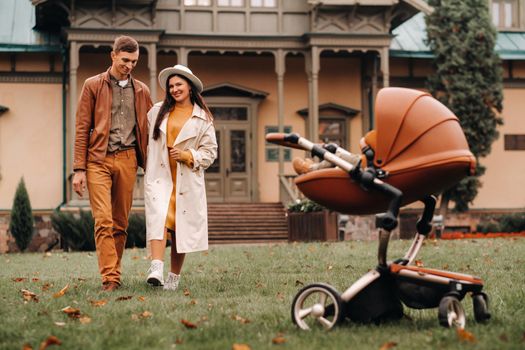 Father and mother on a walk with a child in a stroller in the autumn Park on the background of the estate. A family walks through the Golden autumn nature Park.