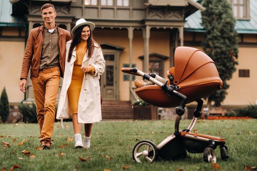 Father and mother on a walk with a child in a stroller in the autumn Park on the background of the estate. A family walks through the Golden autumn nature Park.