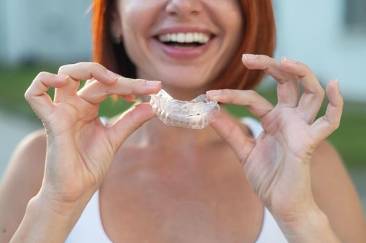 Red-haired Caucasian woman holding transparent mouthguards for bite correction outdoors. A girl with a beautiful snow-white smile uses silicone braces.