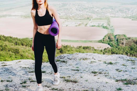 A girl athlete with a purple yoga mat under her armpit. A beautiful athlete in the mountains.