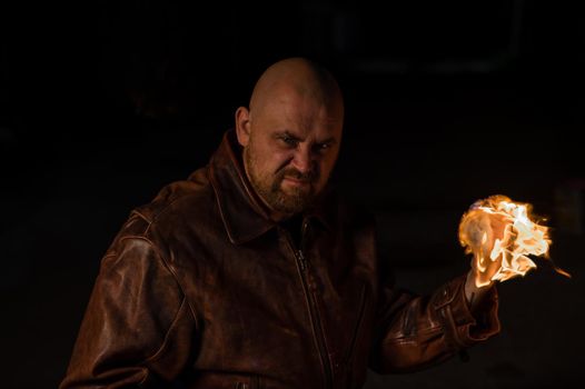 A bald man in a leather jacket holds a burning ball in his hand